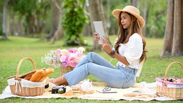 Attraktive asiatische junge Frau in einem lässigen Outfit mit Strohhut liest etwas auf dem Tablet-Bildschirm, während sie in einem schönen Picknick auf der grünen Wiese sitzt. - Foto, Bild