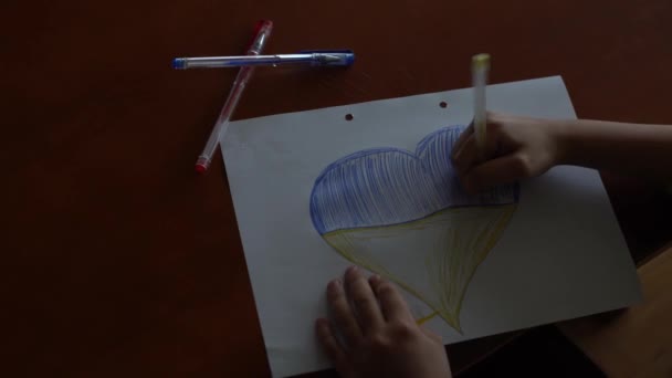Las manos de un niño dibujan un icono en forma de corazón con la imagen de la Bandera Nacional de Ucrania. Vista desde arriba. Niños contra la guerra. Niños dibujando por la paz en Ucrania - Metraje, vídeo