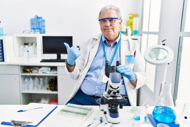 Älterer kaukasischer Mann, der im wissenschaftlichen Labor arbeitet und mit erhobenem Daumen nach hinten zeigt, lächelt selbstbewusst  - Foto, Bild
