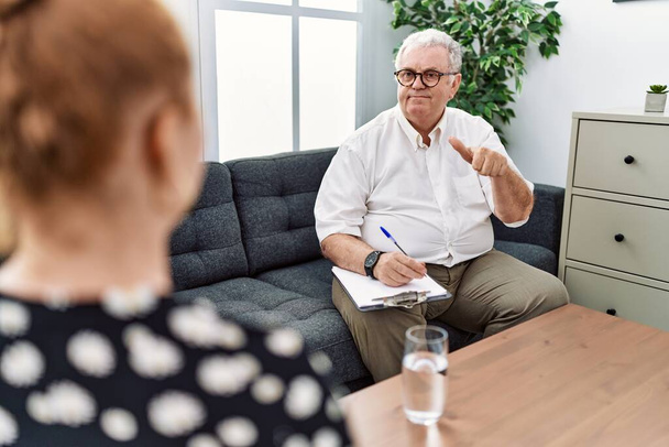 Senior psychologist man at consultation office pointing to the back behind with hand and thumbs up, smiling confident  - Photo, Image