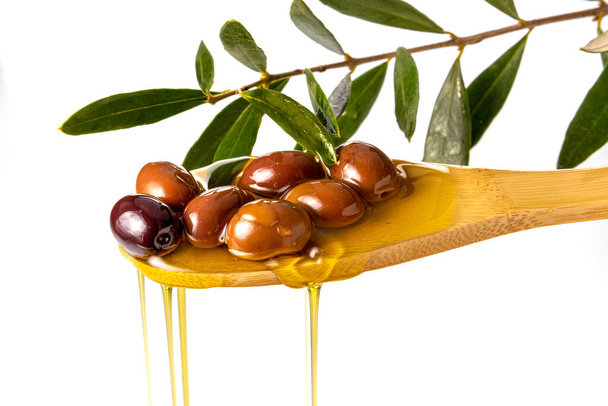 Extra vergine olijfolie druppelend van olijven op houten lepel met olijfblad tak, geïsoleerd op wit - Foto, afbeelding