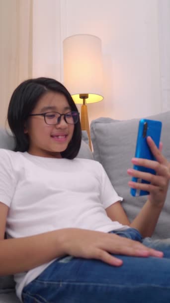 Šťastná asijská dívka sedí na pohovce v obývacím pokoji mluvit s rodičem na mobilním telefonu, usmívající se roztomilá dívka použít video hovor chytrým telefonem mluvit s rodinou. přes WiFi internet. - Záběry, video