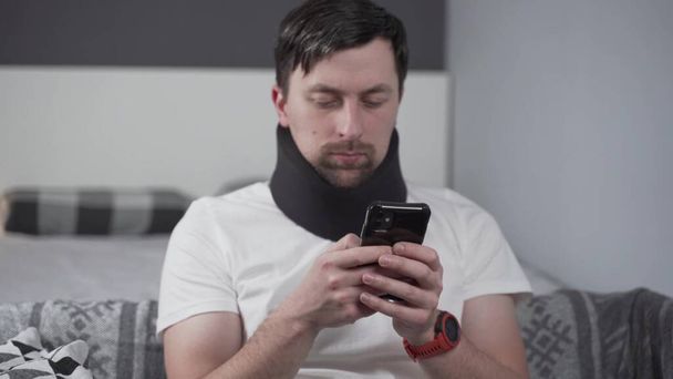 Mladý muž po autonehodě, který trpí doma. Muž v pěnovém krčním límci čte zprávu na mobilu a cítí bolest v krku. Chlápek v krku rovnátka na podporu krku doma na gauči - Fotografie, Obrázek