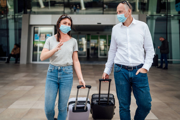 Пара друзей в защитных масках болтают, выходя из аэропорта со своим багажом - Фото, изображение