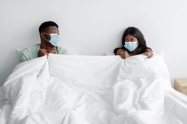 Droevige ongelukkige jonge zwarte zieke vrouw en echtgenoot in beschermende maskers op sociale afstand in bed onder witte deken - Foto, afbeelding