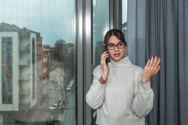 Молодая больная женщина в карантине самоизоляции стоит в своей квартире и смотрит в окно, разговаривая по телефону со своим парнем на улице. Концепция здравоохранения - Фото, изображение