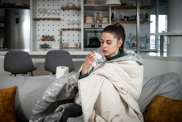 Больной молодой женщины, сидящей под одеялом на диване и чихающей бумагой дома. Женщина, сморкающаяся, кашляющая или чихающая в тканях дома, страдающая гриппом. Простуда и лихорадка. - Фото, изображение