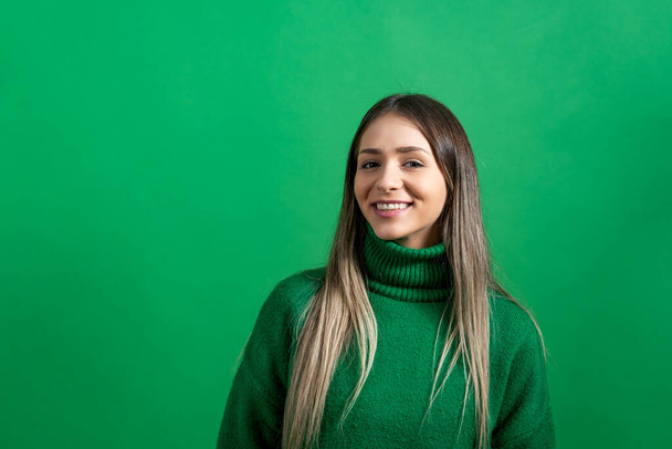 Portrait of young beautiful confident happy woman or teenage girl standing and smiling. Positive female in green sweater standing against green not isolated background. - Photo, Image
