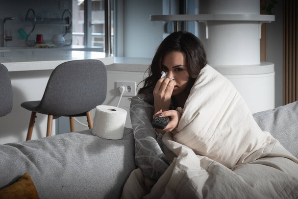 Молода хвора жінка, сидячи вдома на дивані, загорнута в ковдру, відчуває себе виснаженою, дивлячись телевізор, коли вона страждає від застуди. Невдаха жінка має грип і головний біль, що перемикає телеканали
 - Фото, зображення
