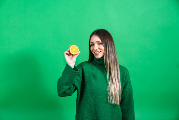 Portrait of young beautiful confident happy woman or teenage girl standing holding orange fruit and smiling. Positive female in green sweater standing against green not isolated background. - Photo, Image