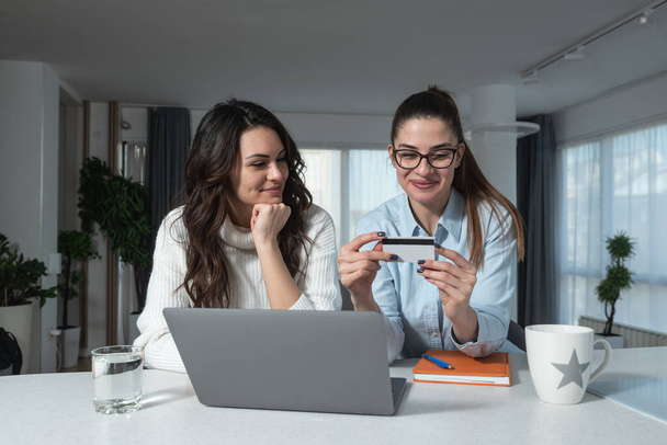 Two happy carefree young women business partners and friends sitting at kitchen counter are happy because they have just tried online shopping with credit card on their new internet store website - Photo, Image