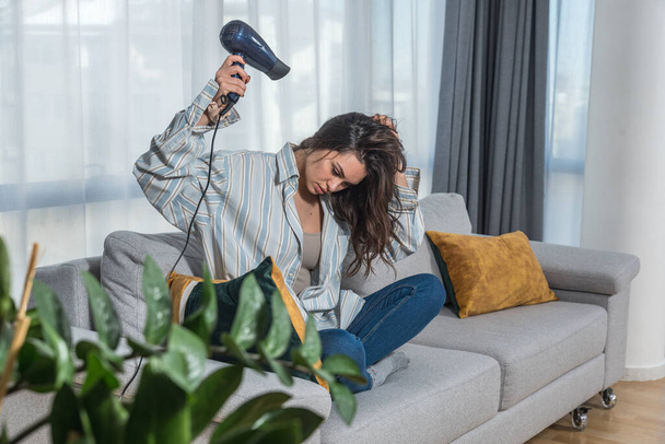 Young beautiful brunette woman drying her long hair at home with hairdryer. Female after showering preparing herself for bed after hard day at work. - Foto, afbeelding