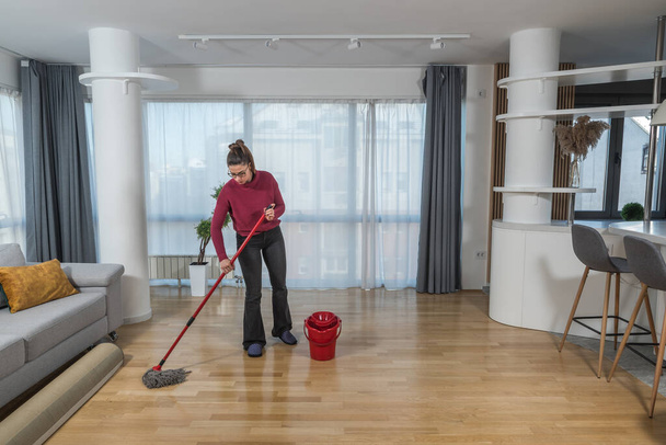 Νεαρή γυναίκα καθαρίζει το πάτωμα του διαμερίσματός της με σφουγγαρίστρα και υγρό σκούπισμα. Γυναίκα κάνει οικιακές εργασίες και τη διατήρηση της υγιεινής του καθαρού σπιτιού για την απομάκρυνση της σκόνης και βρωμιά. - Φωτογραφία, εικόνα