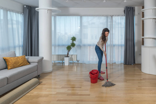 Young woman cleaning her apartment floor with mop and wet wipe. Female doing housework and maintaining hygiene of clean home to remove dust and dirt. - Valokuva, kuva