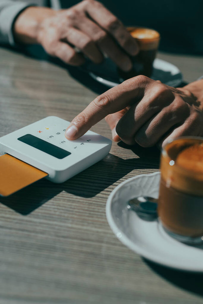 un joven, sentado en una mesa de un café en la acera, inserta el PIN de su tarjeta de crédito en el teclado de un terminal de pago inalámbrico, para pagar la factura - Foto, Imagen