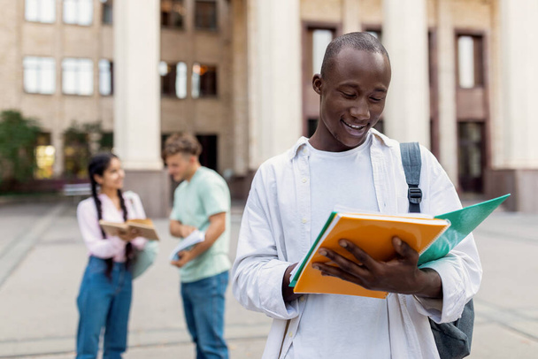 Черный студент университета держит тетрадь и читает, стоит на улице со своими одноклассниками на заднем плане - Фото, изображение