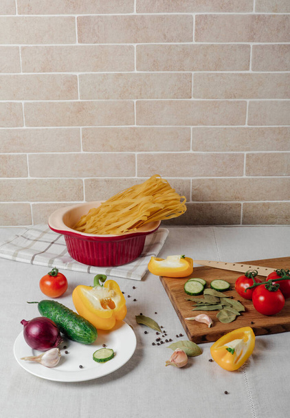 Овощи и сырая паста на столе. Черри помидоры, желтый перец, огурец, красный лук и чеснок на разделочной доске с ножом. Ингредиенты для блюда - Фото, изображение