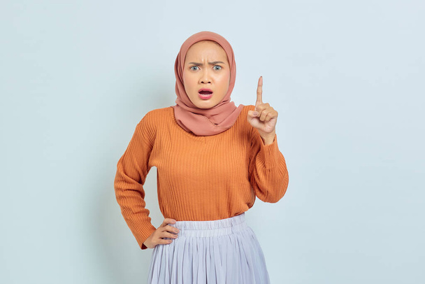 Surprised young Asian Muslim woman in brown sweater pointing to copy space with finger isolated over white background. Muslim lifestyle concept - Photo, image
