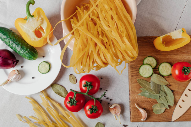 Vegetables and raw pasta on a table. Cherry tomatoes, yellow bell pepper, cucumber, red onion and garlic on the cutting board with knife. Top view - Foto, afbeelding