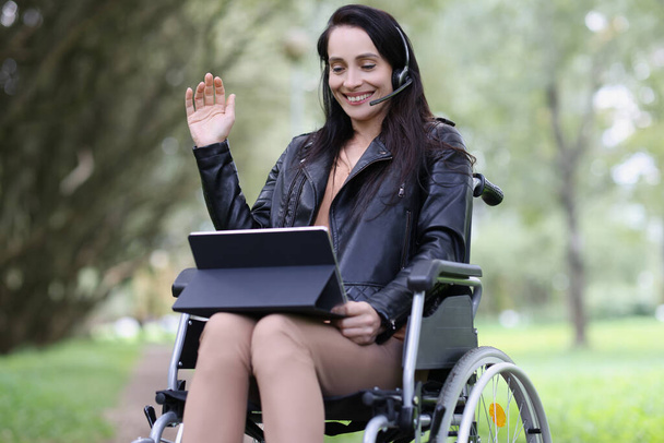 Glimlachende vrouw in hoofdtelefoon en laptop in rolstoel in park - Foto, afbeelding