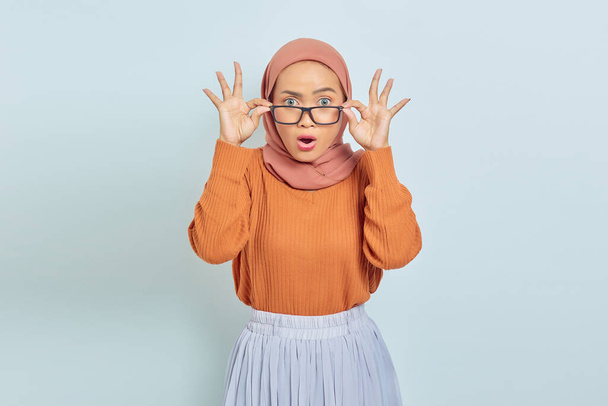 Portrait of surprised young Asian Muslim woman in brown sweater holding glasses and looking at camera isolated over white background. Muslim lifestyle concept - Foto, afbeelding