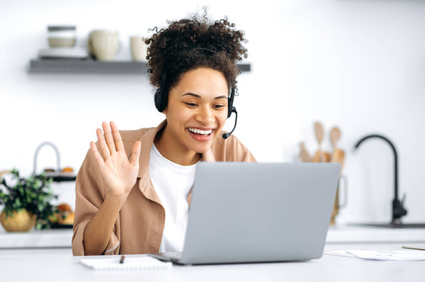 Positive successful african american curly-haired girl, in casual clothes, working or studying from home, using laptop and headset, talking on video conference, greetings, smiling friendly - Photo, Image