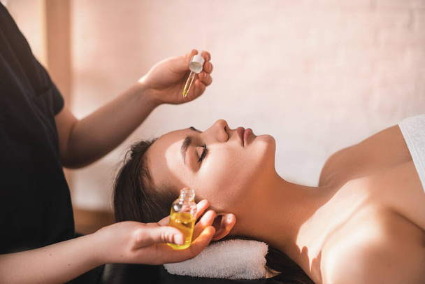 Una hermosa joven recibe un masaje en el spa. El masajista gotea aceite de masaje en la cara. El concepto de cuidado del spa - Foto, imagen
