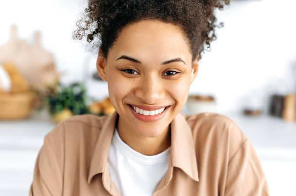 Portrait en gros plan d'une jolie jeune fille afro-américaine joyeuse aux cheveux bouclés et aux taches de rousseur, avec un sourire blanc comme neige, portant une chemise décontractée, regardant la caméra, souriant joyeusement - Photo, image