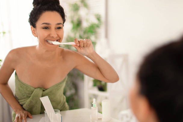 Belle jeune femme avec un beau sourire portant serviette après le bain, brossant les dents avec une brosse à dents près du miroir à la maison - Photo, image