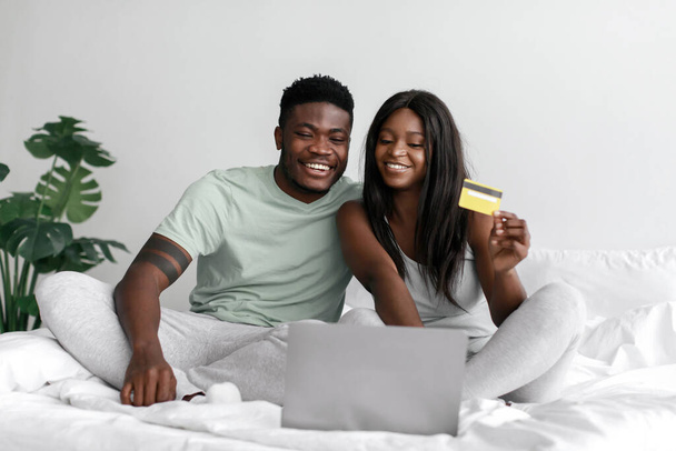 Wesoły młody afrykański mąż i żona patrząc na laptopa i pokazując kartę kredytową, zakupy online - Zdjęcie, obraz