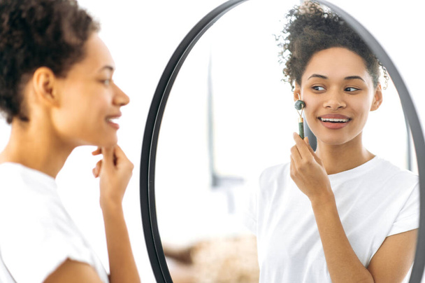 Menina bonita afro-americana em t-shirt branca, fica na frente de um espelho em casa, faz sua rotina de beleza matinal, cuida de sua pele facial, usa um rolo de pedra facial, evita rugas, sorriso - Foto, Imagem