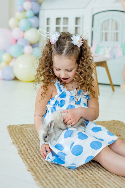 A child girl in bunny ears holding a cute gray rabbit looking at camera in a petting zoo or a farm. Easter bunny in kindergarten for pets. - Foto, Bild