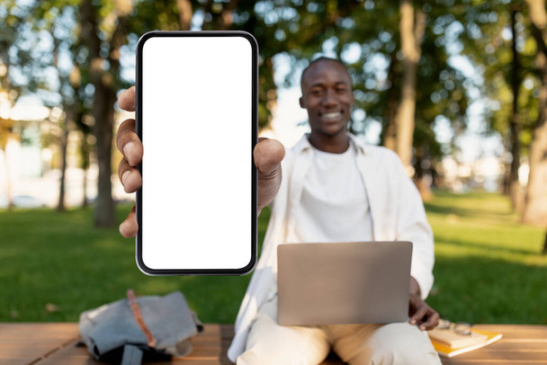 Africain américain étudiant gars démontrant grand smartphone vierge tout en étudiant à l'extérieur avec ordinateur portable, maquette - Photo, image