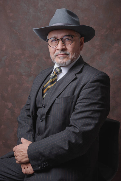 Un caballero maduro posando para la cámara en un traje y sombrero de rayas marrones oscuros - Foto, Imagen