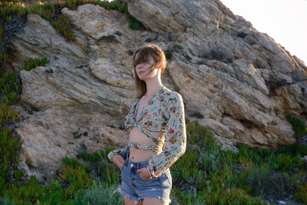 Retrato al aire libre de mujer joven de moda en top de cultivo y pantalones cortos de mezclilla con formación de roca en el fondo - Foto, imagen