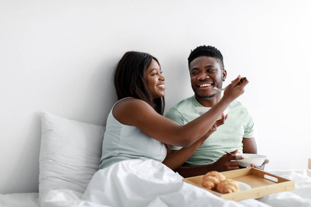 Cheerful happy pretty young african american female feeding male oatmeal and have fun in bed in white bedroom - Photo, Image