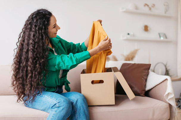 Shopaholic mujer sosteniendo pieza de ropa desembalaje caja en casa - Foto, Imagen