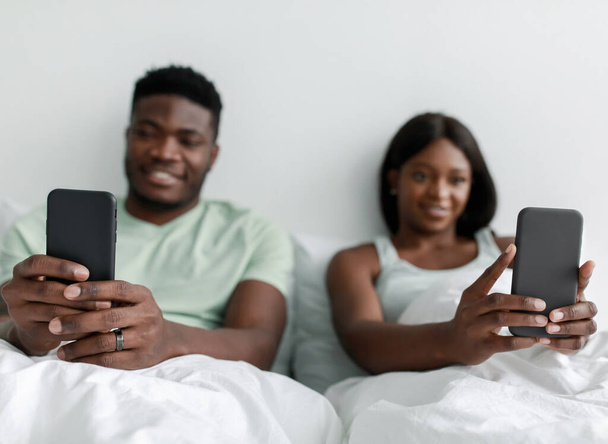 Улыбающиеся молодые африканские муж и жена, печатающие на смартфонах, сидят на кровати в спальне - Фото, изображение