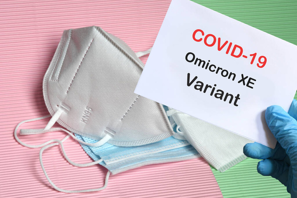 Doctor's hand in blue glove with white paper and text Covid-19 Omicron XE Variant with various protection masks on background. COVID-19 Omicron XE Variant strain protection concept. - Φωτογραφία, εικόνα
