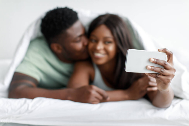 Улыбающийся афроамериканец тысячелетия целует женщину, женщина делает селфи на смартфоне на белой кровати - Фото, изображение