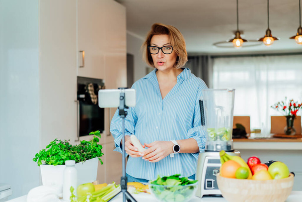 Middle age women is blogging for her wellness lifestyle channel about healthy balanced living in the kitchen of her home. Creating video content for social media with phone camera on a tripod. - Foto, Imagen