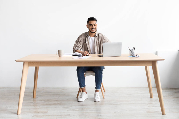 Uśmiechnięty Arab za pomocą laptopa siedzi przy biurku w biurze - Zdjęcie, obraz