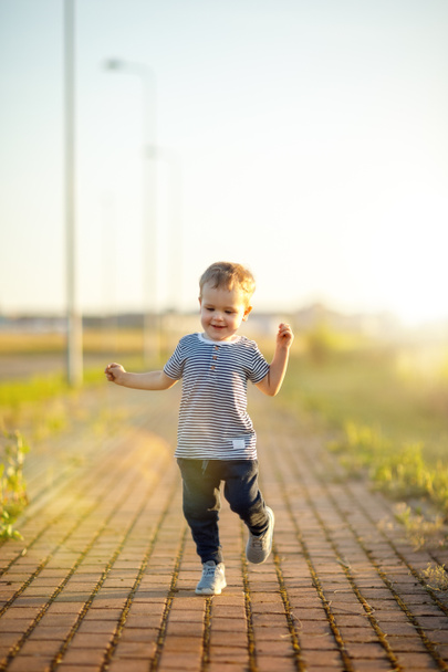 Веселый маленький мальчик в полосатой рубашке бежит по тротуару. Солнечный летний вечер, подсветка. - Фото, изображение