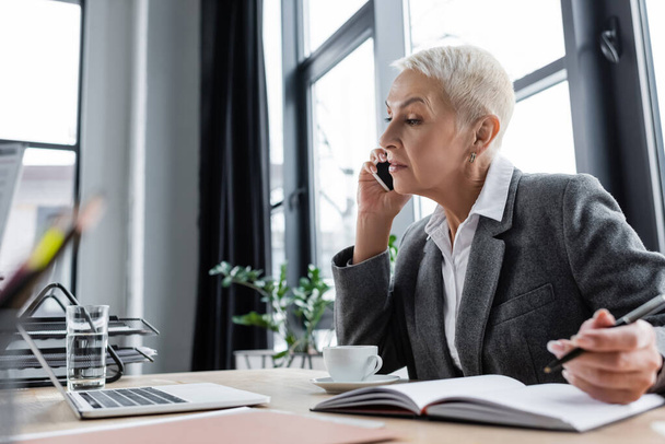 mujer de negocios hablando en el teléfono inteligente mientras mira el portátil cerca de portátil - Foto, Imagen