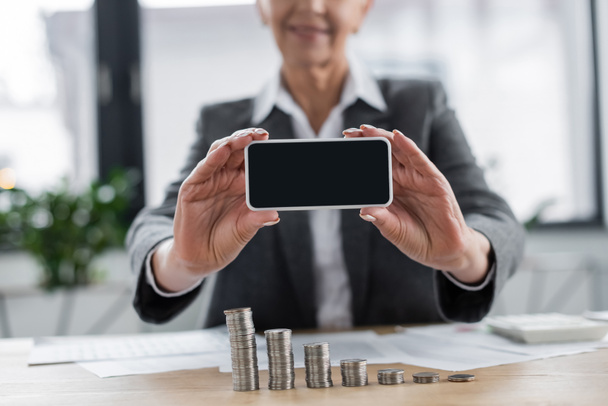 обрезанный вид размытого банкира, держащего мобильный телефон с пустым экраном рядом со стопками серебряных монет - Фото, изображение