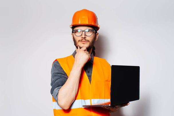 Travailleur de la construction réfléchi avec ordinateur portable dans les mains sur fond blanc. - Photo, image