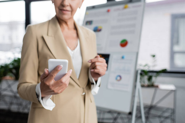 partial view of banker holding smartphone near flipchart on blurred background - Photo, Image