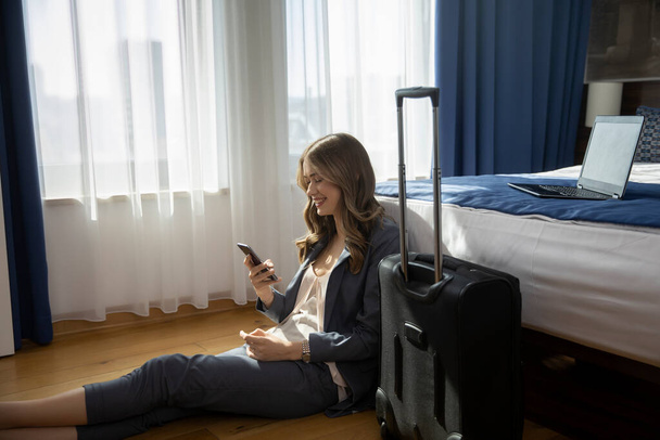 Flight attendant relaxing in a hotel room using her smartphone sitting on a floor - Zdjęcie, obraz