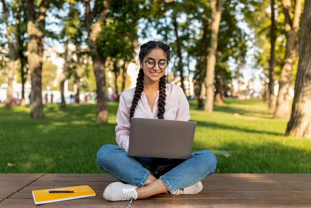 Concepto de aprendizaje electrónico. Estudiante india con portátil sentada en el banco y viendo webinar educativo al aire libre - Foto, Imagen