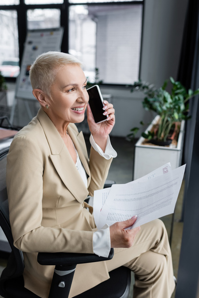 cheerful banker sitting in office with documents and listening voice message  - Photo, Image